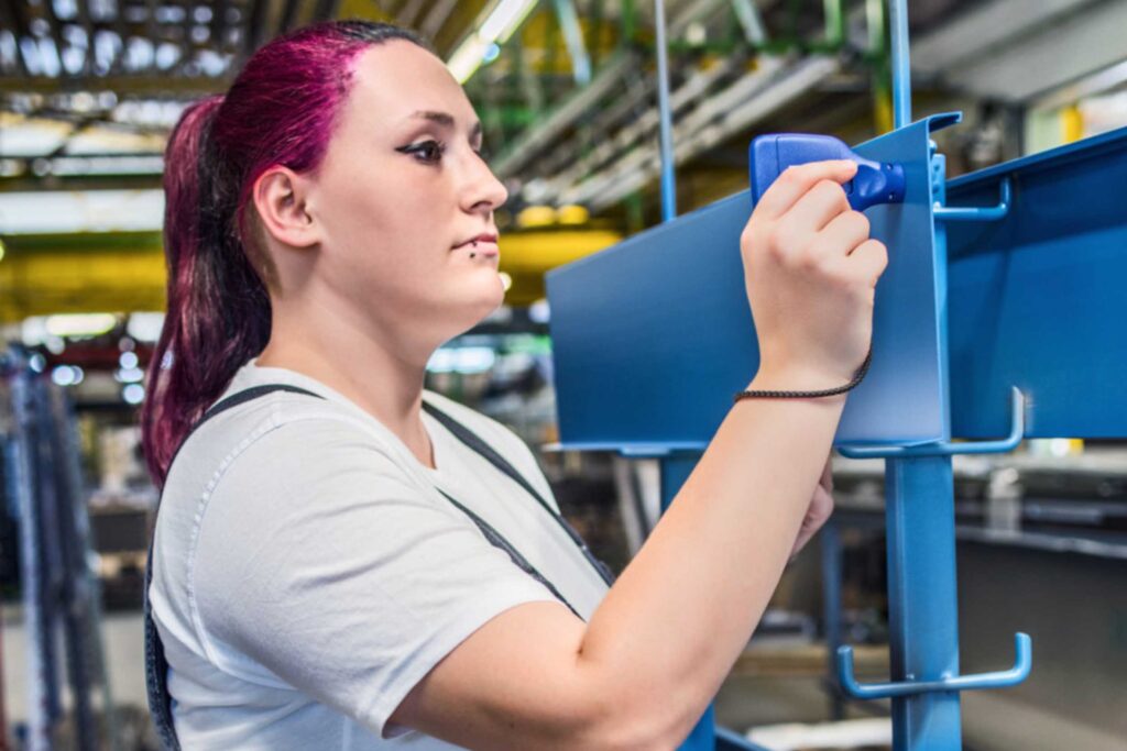 Frau mit lila Haaren und einer blauen Latzhose arbeitet an einem Stahlgerüst